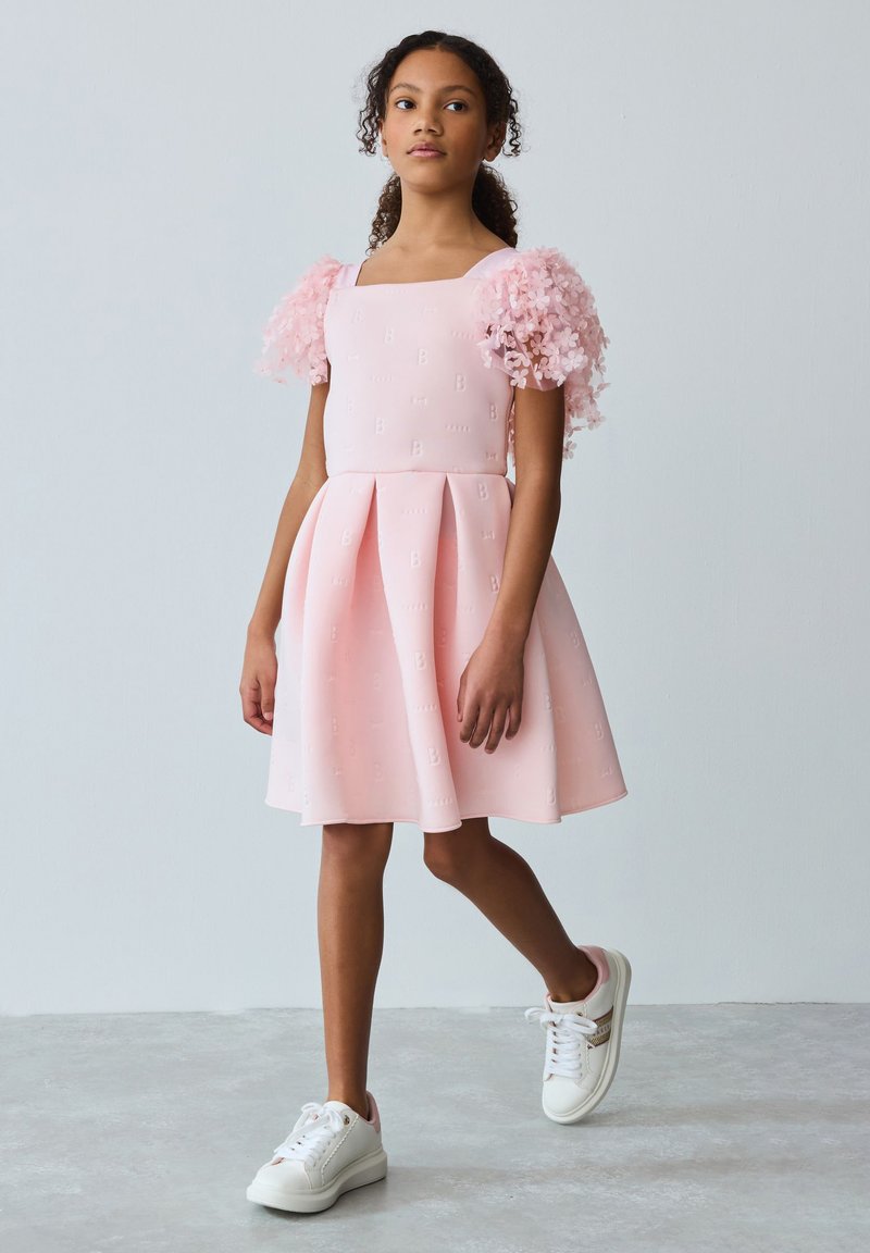 Tyttö kävelee vaaleanharmaata, yksiväristä taustaa vasten pukeutuneena vaaleanpunaiseen mekkoon, jossa on kukalliset, tylliset hihat, ja valkoiset lenkkarit.