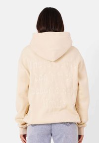 Felpa color crema con una grande citazione ricamata sul retro, caratterizzata da una vestibilità comoda, cappuccio regolabile e polsini a costine.