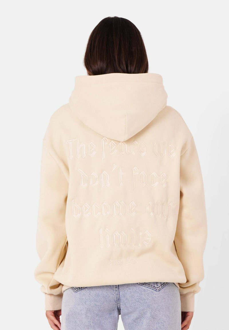 Felpa color crema con una grande citazione ricamata sul retro, caratterizzata da una vestibilità comoda, cappuccio regolabile e polsini a costine.