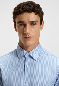 Camicia a righe blu chiaro con colletto strutturato, frontale con bottoni e texture del tessuto liscia. Sottili righe verticali di diverse tonalità di blu.