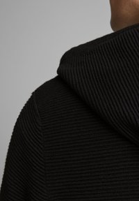 Černá pletená mikina s kapucí s vertikální žebrovanou strukturou, kapucí se stahovací šňůrkou a volným střihem, zobrazena zezadu s měkkým vzhledem.