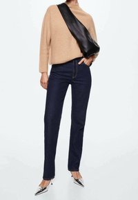 Personne portant un pull beige, un jean bleu foncé, des talons métalliques pointus et un grand sac bandoulière en cuir noir, debout devant un fond uni.