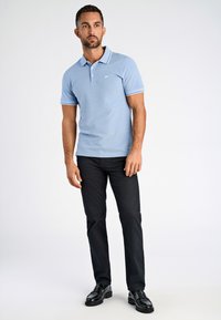 Camisa polo azul claro con contraste blanco en las mangas y el cuello, combinada con pantalones negros y zapatos negros. Logo minimalista en el pecho.