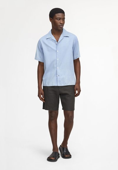 Mann steht und trägt ein hellblaues kurzärmeliges Hemd mit Knöpfen, schwarze Shorts und schwarze offene Sandalen mit Riemen vor weißem Hintergrund.