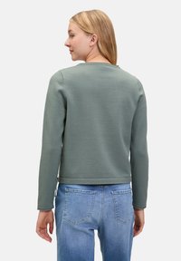 Grüner, langärmeliger Sweatshirt mit rundem Ausschnitt und geripptem Saum, aus einem weichen Material gefertigt. Getragen mit hellblauen Jeans.