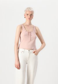 Light pink ribbed tank top with a scoop neckline, decorative trim, button closure at the front, and a cropped fit paired with white wide-leg pants.