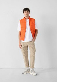 Oranje gewatteerde mouwloze vest over een wit t-shirt, gecombineerd met beige cargobroek en grijze sneakers. Eenvoudig ontwerp met een gladde textuur.