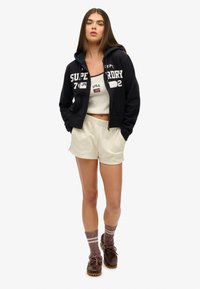 Sudadera negra con cremallera y texto blanco, top corto crema, pantalones cortos beige, zapatos marrones con cordones y calcetas de punto con rayas rosas.