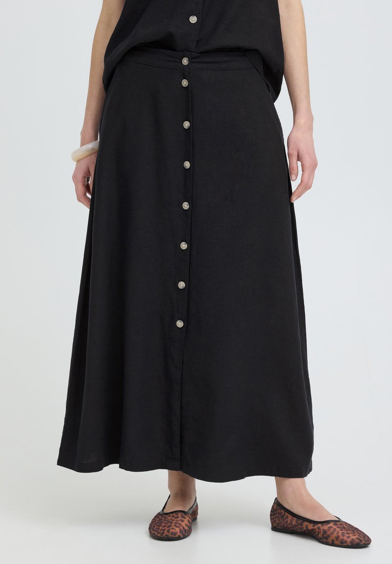 Femme portant une longue jupe noire boutonnée, un haut noir, des ballerines imprimé léopard et un bracelet blanc au poignet gauche.