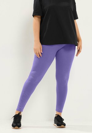 Kvinde iført lyse lilla leggings, sort t-shirt og sorte sneakers, stående mod en hvid baggrund.