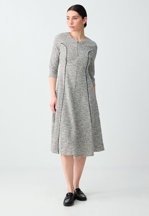 Femme debout portant une robe midi grise avec des détails en passepoil noir, des manches trois-quarts et des chaussures noires sans lacets, regardant sur le côté.