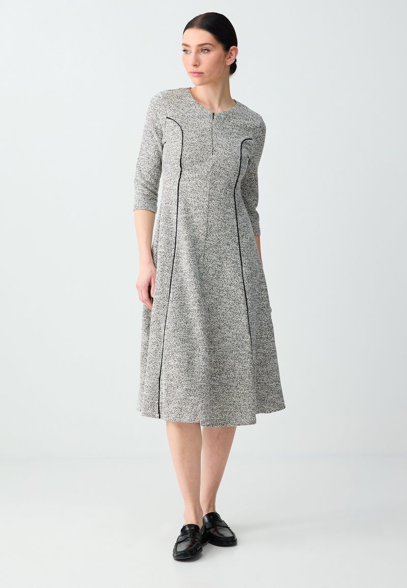 Femme debout portant une robe midi grise avec des détails en passepoil noir, des manches trois-quarts et des chaussures noires sans lacets, regardant sur le côté.