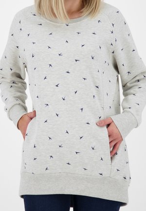 Donna che indossa una felpa grigio chiaro con piccole stampe di uccelli blu scuro, mani nelle tasche anteriori, abbinata a jeans blu scuro.