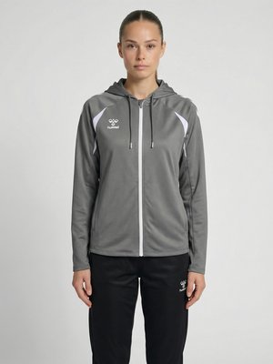 Femme debout face à l'avant, portant une veste à capuche grise zippée et un pantalon noir avec des logos blancs sur un fond clair uni.