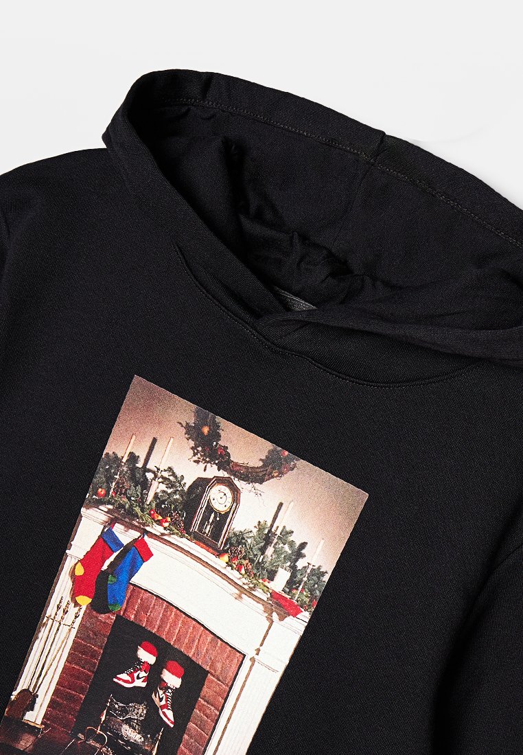 Sweatshirt à capuche noir avec un motif imprimé représentant une cheminée décorée pour les fêtes, avec des chaussettes, des bougies et de la verdure. Tissu doux et décontracté.