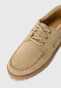 Zapato de ante en color beige con cordones y punta redondeada, costuras en contraste y suela de goma. Presenta ojales en tono dorado y un forro interior suave.