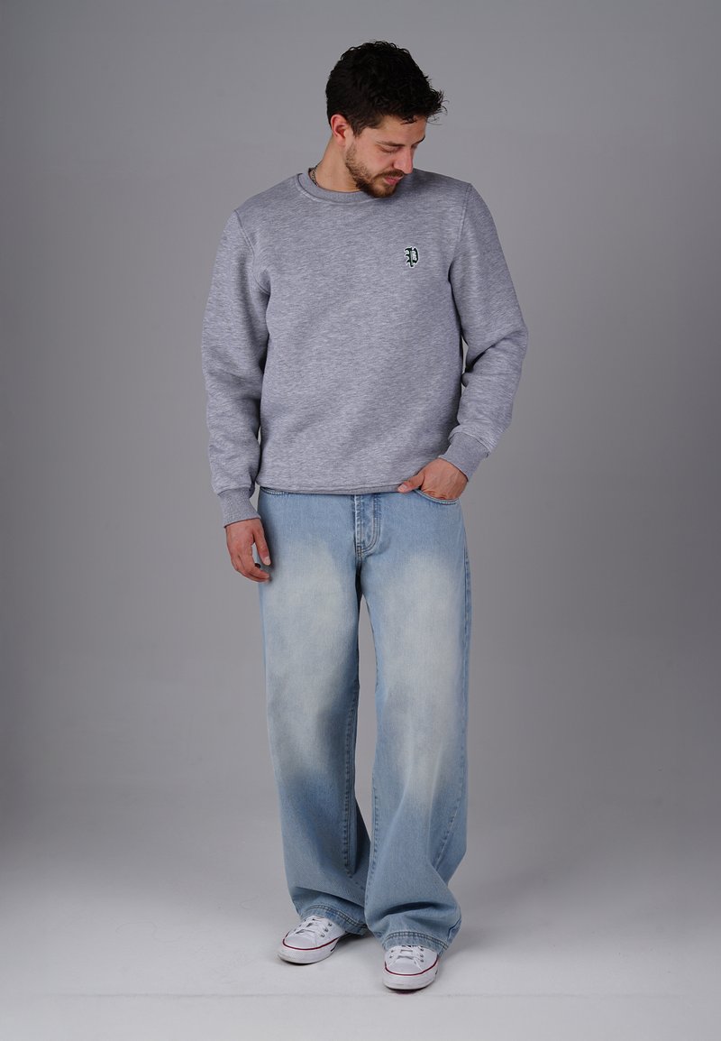 Grauer Sweatshirt mit Rundhalsausschnitt und gesticktem grünen Logo. Hellblaue weit geschnittene Jeans mit ausgewaschenem Finish und weißen Sneakern.