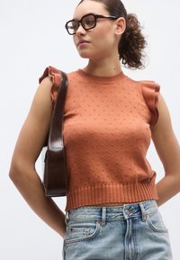 Πλεκτό αμάνικο τοπ σε σκουρόχρωμο πορτοκαλί με υφή, ριμπ τελείωμα και ανάγλυφη λεπτομέρεια στο ώμο, σε συνδυασμό με ανοιχτόχρωμο μπλε τζιν παντελόνι.