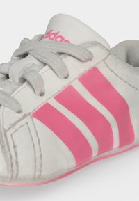 Λευκό παπούτσι Adidas με ροζ ρίγες και κορδόνια, γωνιακά για να δείξει τις πλευρικές και πάνω μπροστινές λεπτομέρειες.