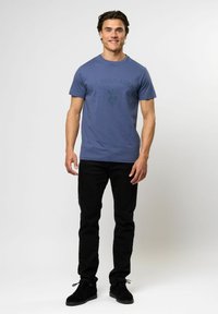 Mėlynas medvilninis marškinėlis su tamsiai mėlynu U.S. Polo Assn. logotipu, derinamas su juodomis kelnėmis ir juodais batais. Modelis stovi ramiai, atsukęs į priekį.