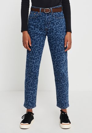 Osoba nosząca niebieskie dżinsy w cętki lamparta, brązowy pasek, czarny top z długim rękawem oraz czarne trampki z białymi podeszwami, stojąca na białej podłodze.