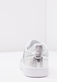Stříbrné metalické tenisky s bílou podrážkou, s hladkou texturou, šněrovacím designem a embosovaným logem "Reebok" na patě.