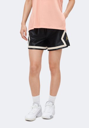 Person iført sorte basketballshorts med hvide kanter, ferskenfarvet ærmeløs top, hvide sokker og hvide sportssko stående mod en ensfarvet baggrund.