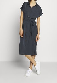 Abito camicia grigio scuro a maniche corte con colletto, bottoni frontali, cintura in vita e due tasche sul petto, abbinato a sneakers bianche.