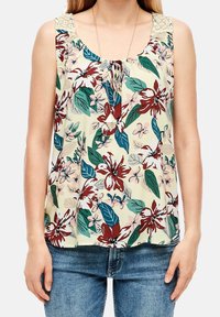Femme portant un haut sans manches à motif floral avec des détails en dentelle aux épaules et un jean bleu, accessoirisée d’un long collier pendentif.