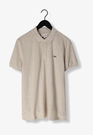 Polo beige à manches courtes avec col et deux boutons, arborant un petit logo crocodile vert sur la poitrine gauche, suspendu à un cintre noir.