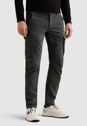 Muž v tmavosivých cargo nohaviciach s viacerými vreckami a bielych teniskách s čiernymi podrážkami, stojaci pred jednoduchým pozadím.