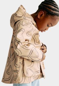 Beige vandtæt jakke med hætte, der har et sort abstrakt swirl-mønster, frontknaplukning og blødt foder.