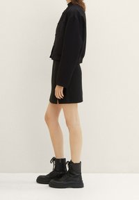 Femme portant une veste noire texturée, une mini-jupe assortie avec une fente latérale, et des bottines noires à lacets, sur un fond neutre.
