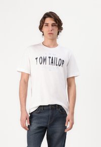 Valkoinen puuvillapaita, jossa on tummansinisellä "TOM TAILOR" -grafiikka, lyhyet hihat, pyöreä kaula-aukko, yhdistettynä tummansineihin farkkuihin.