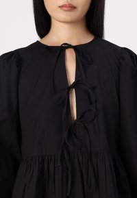 Vestido de algodão preto com uma abertura em forma de olho-chave na frente, fechos amarrados e mangas bufantes com detalhes franzidos na cintura.