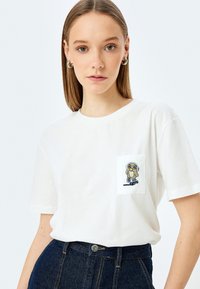 Witte katoenen t-shirt met een zak en een geborduurd cartoonontwerp. Ronde halslijn en korte mouwen; gladde stoftextuur.