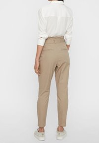 Beige tailored pants with a high waist, tapered legs, and back pockets. Paired with a white collared shirt and light-colored sneakers.