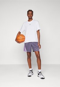 Witte T-shirt met korte mouwen en zwart logo, paarse basketbalshorts met gradient accenten, witte sokken en zwart-witte sneakers, terwijl hij een oranje basketbal vasthoudt.