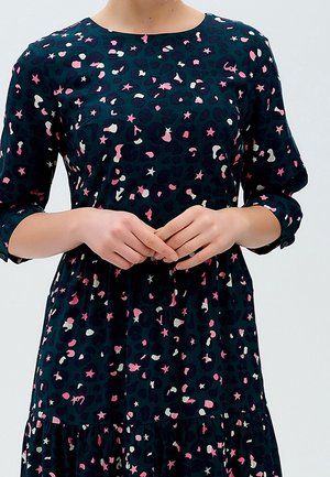 Frau trägt ein dunkles Kleid mit rosa, weißen und schwarzen Stern- und abstrakten Mustern, hält die Hände locker vor sich.