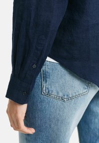 Navyblauw linnen overhemd met knoopmanchetten, gecombineerd met lichtblauwe spijkerbroek met een puntige achterzakontwerp en zichtbare stiksels.