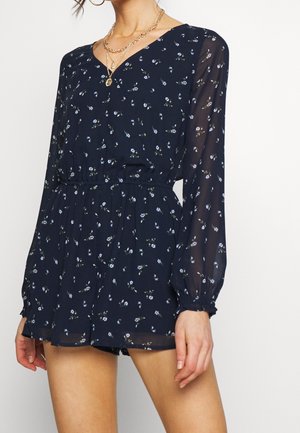 Vrouw draagt een marineblauwe, met bloemenmotief, lange mouwen romper met elastische taille en gelaagde gouden kettingen, staand tegen een effen witte achtergrond.