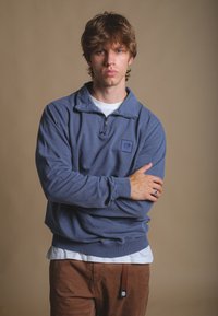 Modrý fleecový svetr s polovičním zipem, ribovanými manžetami a logem. Nositý přes bílý tričko, doplněný hnědými kalhotami. Hladká textura.