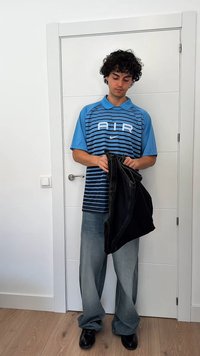 Jovem com cabelo encaracolado segura umas calças pretas, vestindo uma camiseta azul riscada da Nike com a inscrição "AIR", calças largas e sapatos pretos, de pé junto a uma porta branca.
