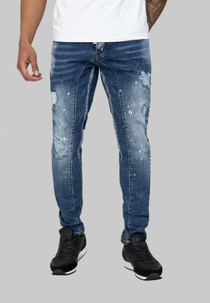 Jean en denim bleu déchiré avec des détails déchirés et des éclaboussures de peinture colorées. Design ajusté avec poches standard et légèrement extensible.