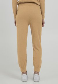 Beige joggerbukser fremstillet af blødt strikmateriale, med en tapered benpasform, elastisk talje og ribbede manchetter. Bæres med hvide sneakers.