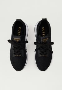 Melnas sporta kurpes, kas izgatavotas no struktūras adījuma materiāla ar melnām saitēm, zelta logotipa emblēmu un baltu gumijas zole.