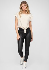 Top corto beige con nodo anteriore, abbinato a leggings neri a vita alta. Modella che indossa sneakers bianche con una texture liscia.
