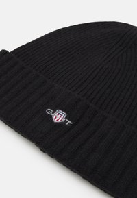 Zwart gebreid beanie-muts met ribbelstructuur, voorzien van een klein geborduurd logo in rood, wit en blauw nabij de onderrand.