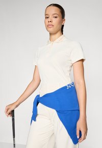 Kremfarget polo skjorte med krage, blå hettegenser bundet rundt livet, beige bukser, holder en svart golfklubbe. Glatte teksturer, enkelt design.