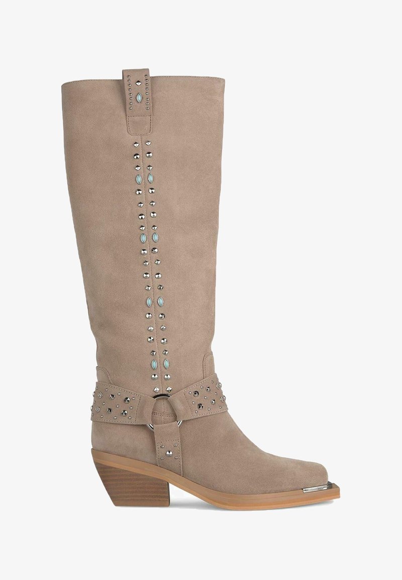 Bottes en suede beige arrivant jusqu'aux genoux avec un bout carré, ornées de clous décoratifs et d'un accent en métal sur la boucle. Design à talon bloc.